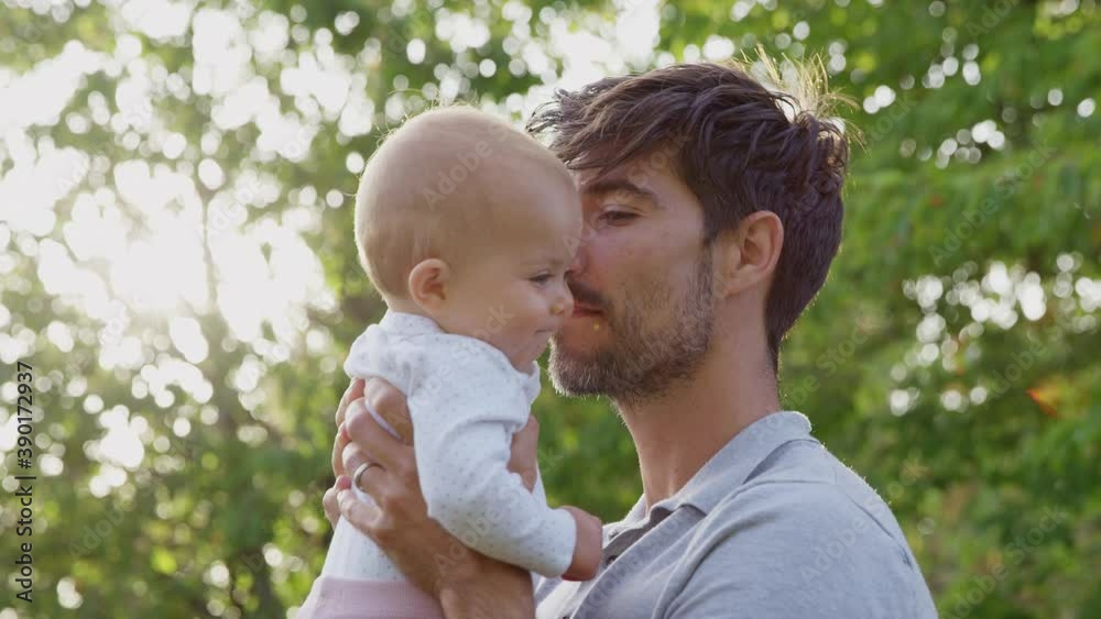 Loving father cuddling and kissing baby daughter as he holds her outdoors in park or garden against flaring sun - shot in slow motion Loving father cuddling and kissing baby daughter as he holds her outdoors in park or garden against flaring sun - shot in slow motion