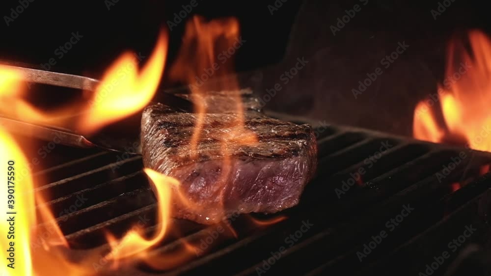 Juicy beef steak is fried on fire coals on iron grill on dark background in flame of smoke. Barbecue in evening, chef turns meat on grill with tongs. cooking dinner in a hot flame.