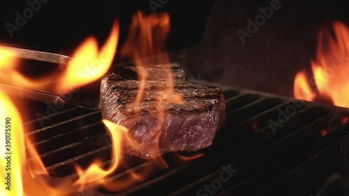 Juicy beef steak is fried on fire coals on iron grill on dark background in flame of smoke. Barbecue in evening, chef turns meat on grill with tongs. cooking dinner in a hot flame.