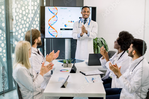 Multiethnic team of doctors applauding to cheerful handsome African male colleague, standing near the digital screen and having successful speech about DNA structure and new medicines