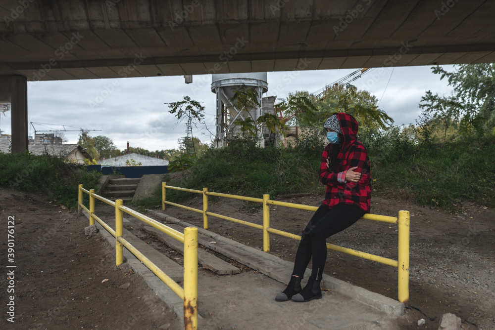 Young depressed homeless girl or woman with hood and protective medicine mask sitting outdoor scared from coronavirus virus or COVID-19 feeling abandoned and anxious selective focus Young depressed homeless girl or woman with hood and protective medicine mask sitting outdoor scared from coronavirus virus or COVID-19 feeling abandoned and anxious selective focus