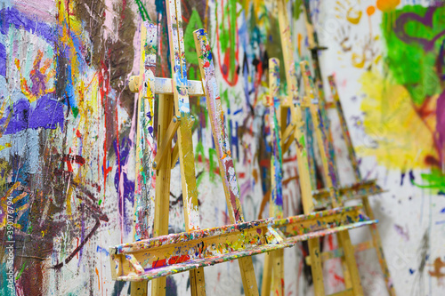 An easel, stained with paint, stands against the background of a painted wall. Art workshop.