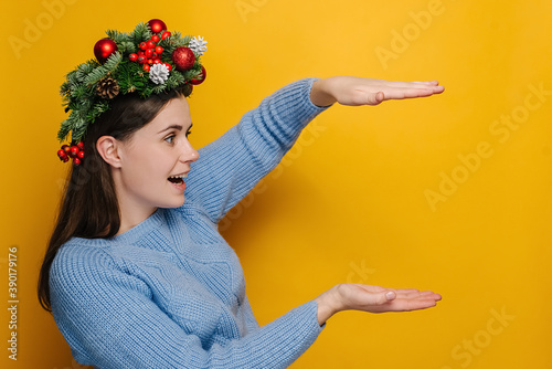 Shocked young woman in Christmas wreath showing something large of big size looking at hands, amazed girl with open mouth surprised with huge discounts gesturing, isolated on yellow studio background