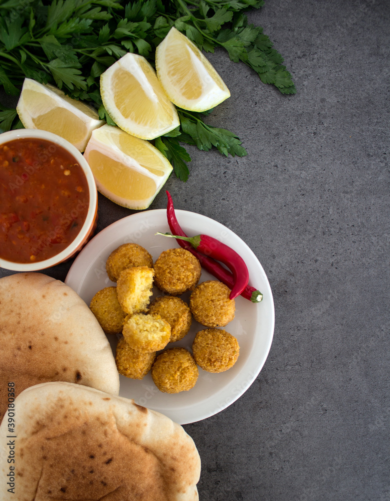 Falafel - authentic food of Israel. Fresh ingredients top view: plate of falafels, pita bread, parsley, harissa sauce, lemon, parsley and red pepper.  Dark grey background with copy space. 