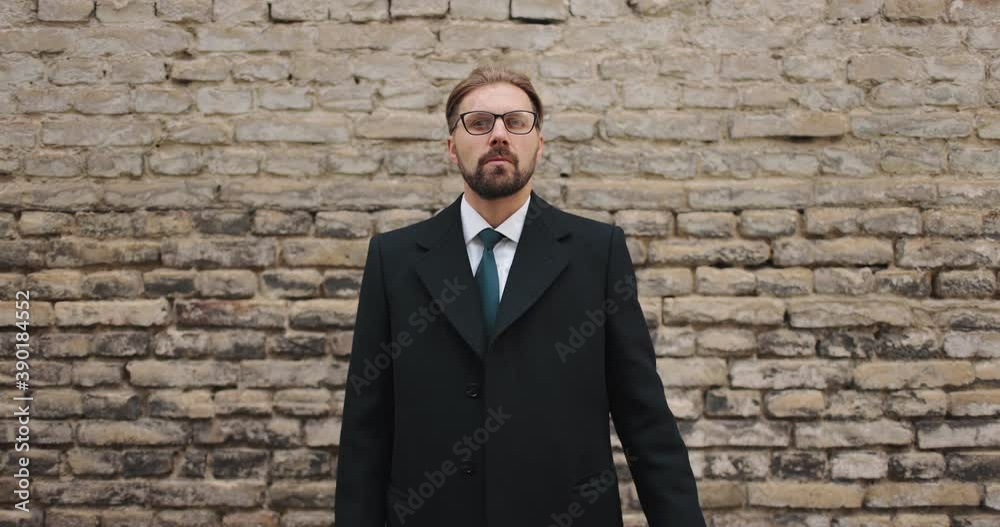 Portrait of handsome bearded man in black coat and suit posing on camera with brick wall behind. Businessman in eyeglasses standing outdoors and looking ahead. Portrait of handsome bearded man in black coat and suit posing on camera with brick wall behind. Businessman in eyeglasses standing outdoors and looking ahead.