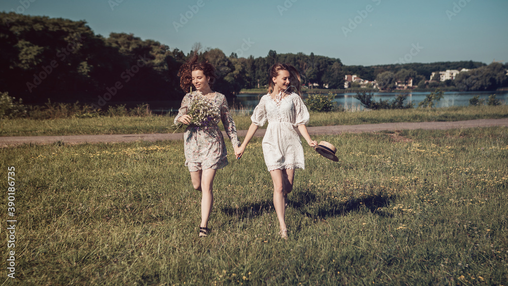 Two ladies running on the grass coast in the sunny morning summer country near the river or lake. Woman dressed in bright dress hold a bouquet of gathered wildflowers, nature background copy space.