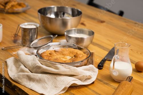 Tasty apple pie on a oaken wood table with a ingredients. Cooking a delicious apple pie on a kitchen in a loft style. Milk, eggs, sugar and a baked pie on a table. Home kitchen.
