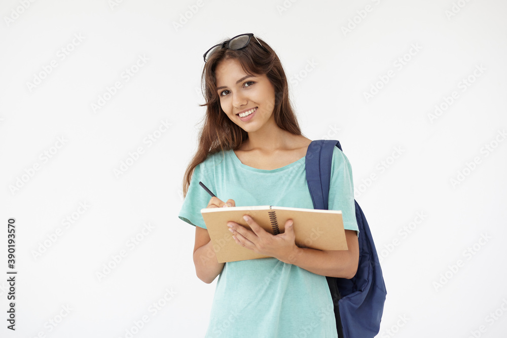 Picture of cheerful positive Hispanic college girl with eyeglasses on her head and bag on one shoulder posing in studio, smiling happily while writing something down in her copybook or diary Picture of cheerful positive Hispanic college girl with eyeglasses on her head and bag on one shoulder posing in studio, smiling happily while writing something down in her copybook or diary