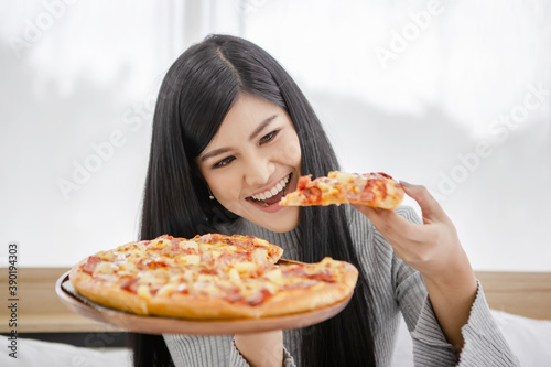 Beautiful happy smiling girl sitting on the bed showing a whole pizza and trying to eat a piece of pizza in her hand for her breakfast	
