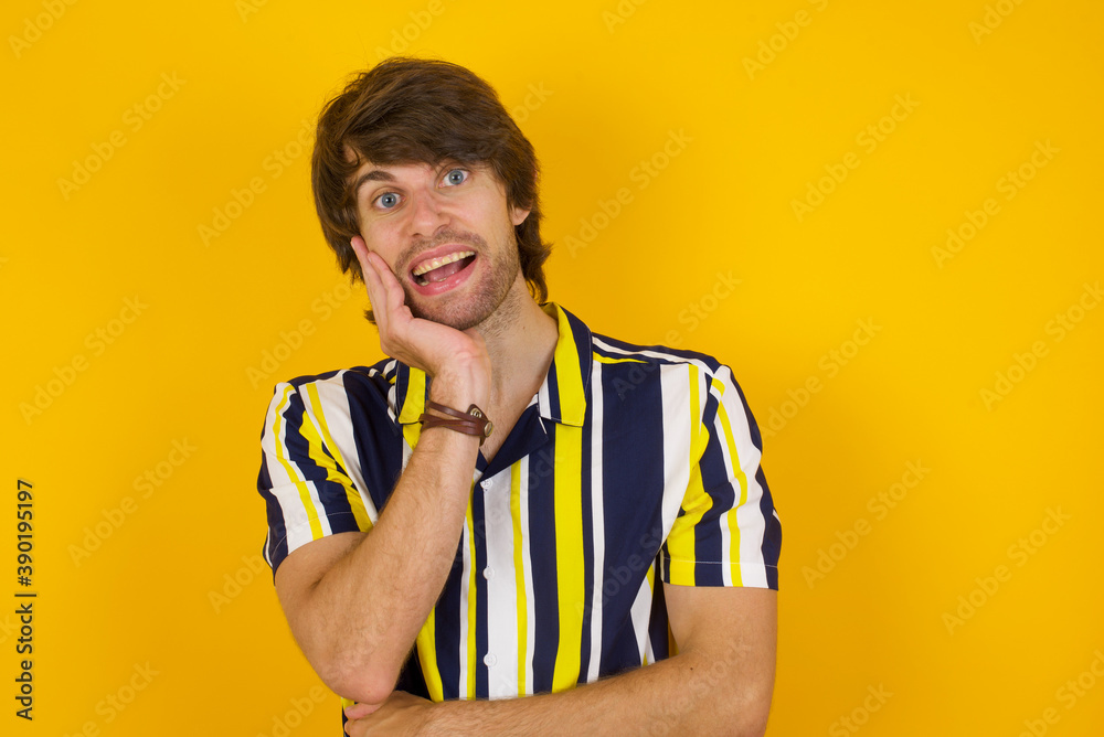 Shocked, astonished Young handsome Caucasian man, wearing stripped shirt  looking surprised in full disbelief wide open mouth with hand near face. Positive emotion facial expression body language.