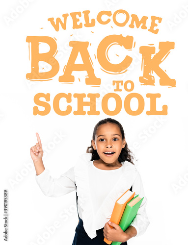 african american schoolgirl pointing with finger at welcome back to school illustration while holding books isolated on white
