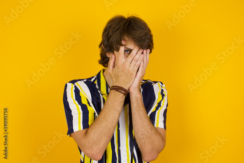 Young handsome Caucasian man, wearing stripped shirt standing against yellow wall covering face with hands and peering out with one eye between fingers. Scared from something or someone.
