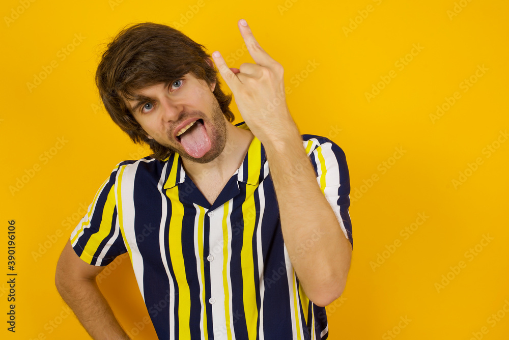 Portrait of a crazy Young handsome Caucasian man, wearing stripped shirt standing against yellow wall showing tongue horns up gesture, expressing excitement of being on concert of band. Portrait of a crazy Young handsome Caucasian man, wearing stripped shirt standing against yellow wall showing tongue horns up gesture, expressing excitement of being on concert of band.