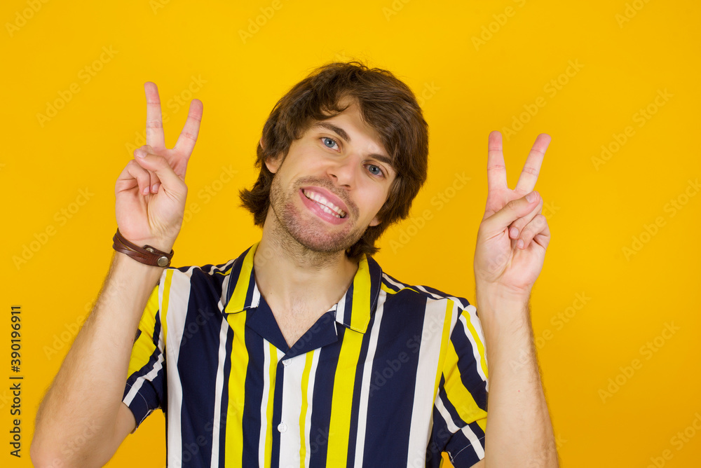 Isolated shot of cheerful Young handsome Caucasian man, wearing stripped shirt standing against yellow wall makes peace or victory sign with both hands, feels cool. Isolated shot of cheerful Young handsome Caucasian man, wearing stripped shirt standing against yellow wall makes peace or victory sign with both hands, feels cool.