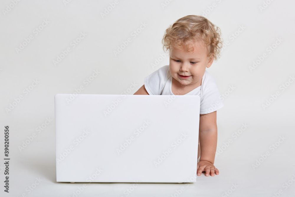 Cute little boy with white laptop computer, looking at screen with interest, child with curious facial expression sitting on floor, wearing t shirt, infant watches cartoons. Cute little boy with white laptop computer, looking at screen with interest, child with curious facial expression sitting on floor, wearing t shirt, infant watches cartoons.
