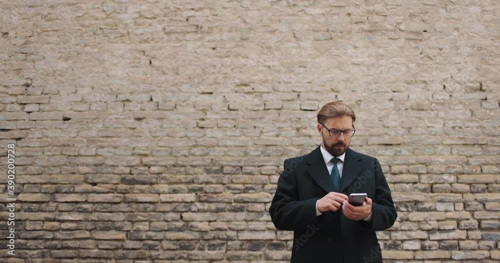 Portrait of handsome bearded businessman in formal outfit making call on mobile while standing outdoors. Background of brick wall. Concept of technology. Portrait of handsome bearded businessman in formal outfit making call on mobile while standing outdoors. Background of brick wall. Concept of technology.