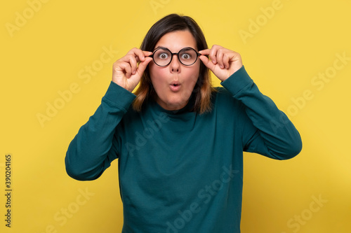 Surprised caucasian woman in glasses looking on interesting information. Studio shot on yellow wall.