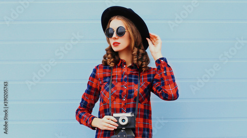 Portrait of young woman photographer with vintage film camera wearing a black round hat over blue background