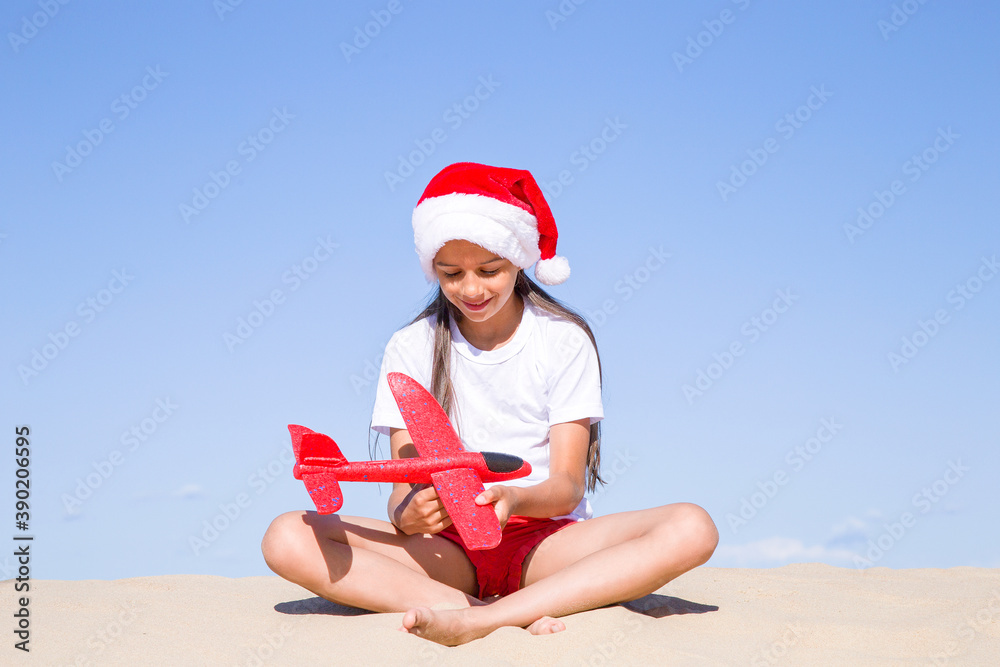 Obraz premium Christmas celebration vacations on a tropical beach: Happy cute little girl wearing a red Santa hat sitting on the sandy beach by the sea and holding a red toy plane on her hands on clear sunny day