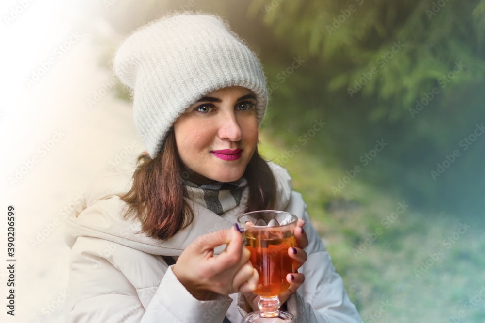 A close up portrait of a young redhead woman dressed in white winter clothes smiling with inspiration and holding a tall glass cup of warming tea while on walk through a mountain forest A close up portrait of a young redhead woman dressed in white winter clothes smiling with inspiration and holding a tall glass cup of warming tea while on walk through a mountain forest