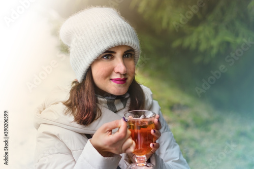 A close up portrait of a young redhead woman dressed in white winter clothes smiling with inspiration and holding a tall glass cup of warming tea while on walk through a mountain forest