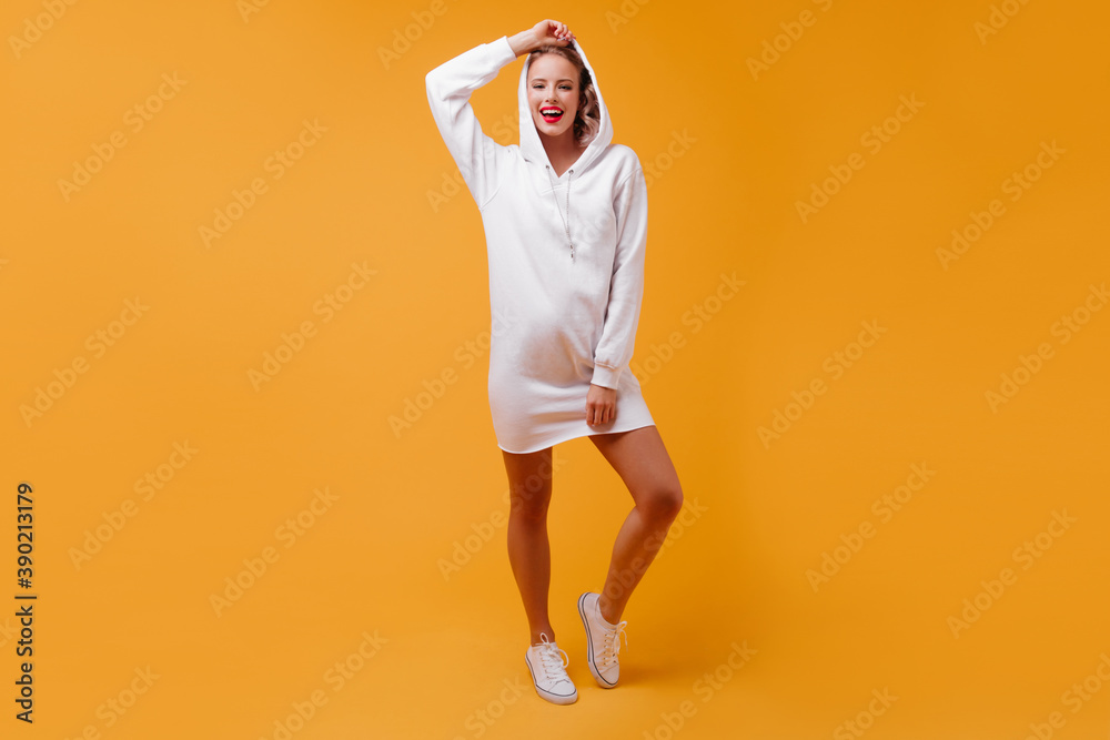 Optimistic girl puts on hood, posing for full-length photo indoors. Pretty student in white outfit smiling cute - obrazy, fototapety, plakaty 