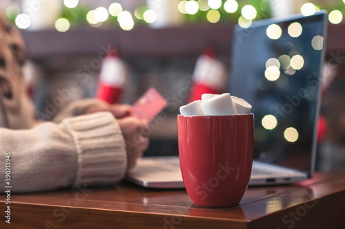 Woman  holding credit card using laptop for making order sitting at table with cup of hot cocoa and marshmallow at christmas fireplace with decoration of light bulbs. Close up.