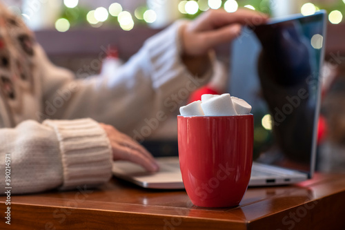 Lonely woman freelancer enjoying with cup of hot cocoa and marshmallow working on laptop near christmas fireplace with decoration of light bulbs.