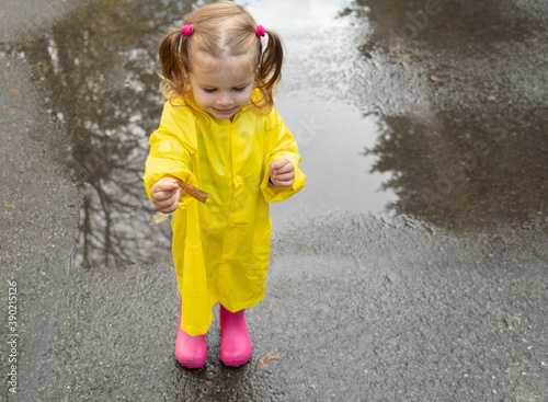 Cute baby girl toddler wearing yellow stylish raincoat pink rubber boots standing in a puddle.