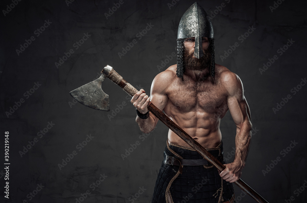 Nord barbarian with naked torso and beard in helmet posing holding his two handed axe in dark studio background. Nord barbarian with naked torso and beard in helmet posing holding his two handed axe in dark studio background.