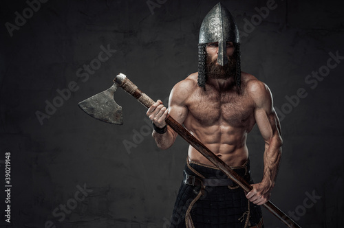 Nord barbarian with naked torso and beard in helmet posing holding his two handed axe in dark studio background.
