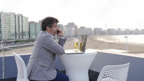 A man working on the rooftop terrace of a bar talking on the phone with the computer on the table and drinking a beer