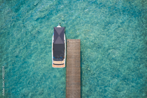 A large white boat with an awning is moored at a wooden pier. Boat with a motor on transparent turquoise water top view. Aerial view of a white yacht at the pier. Copy space. High altitude top view © Berg