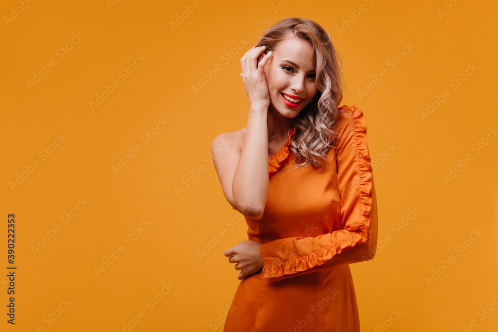 Lovely girl with cute curly hairstyle smiling on yellow background. Studio shot of enchanting white lady in orange dress touching her hair Lovely girl with cute curly hairstyle smiling on yellow background. Studio shot of enchanting white lady in orange dress touching her hair