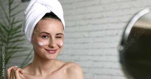 Happy young woman wrapped in bath towel putting cream on face, smiling, looking at mirror and winking to herself. Concept of skincare routine.