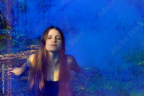 Red-haired woman sitting in a black long dress in a puff of blue smoke in the forest. Mysticism