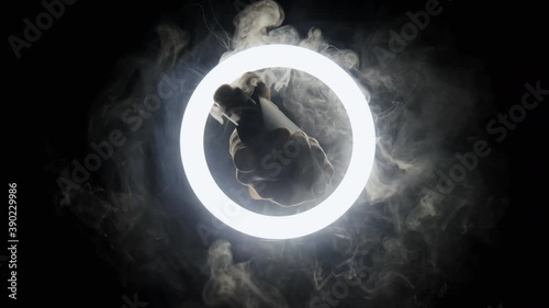 Hand with vape and smoke in slow motion passes through the circle of light on black background. Steam spray from from an electronic cigarette