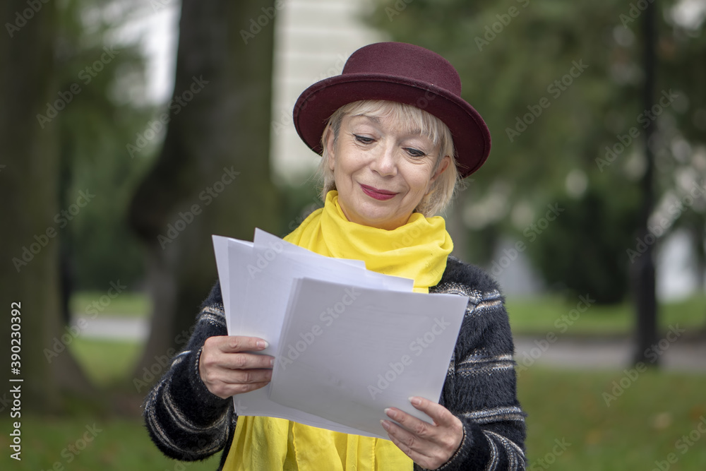 An elderly pensioner 60-65 years old reads documents with text sitting in the Park on a bench. Perhaps she is a teacher and checks the work of students or is preparing to give a lecture.