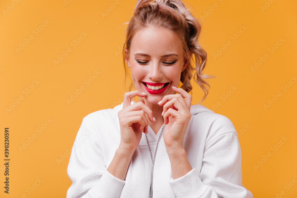 Studio close-up snapshot of girl with bright lips shining from happiness. Cute student in white hoodie closes her eyes and enjoys moment