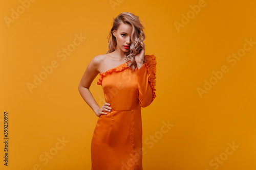 Fashionable caucasian woman in orange dress looking to camera. Confident spectacular blonde girl posing on yellow background