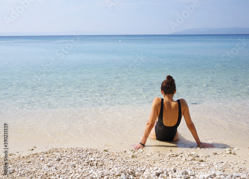 Beautiful young tanned girl in a red bikini sits on the shore of a paradise white sand beach with bruise color in the sea