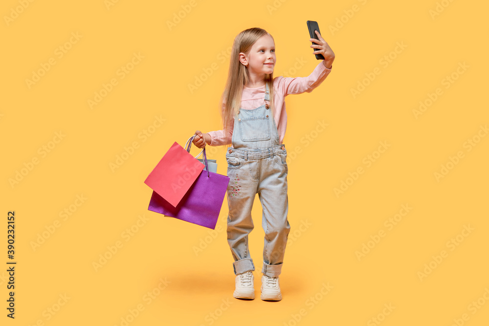 A full-length little girl takes a selfie with purchases in her hands. Yellow isolated background. A full-length little girl takes a selfie with purchases in her hands. Yellow isolated background.