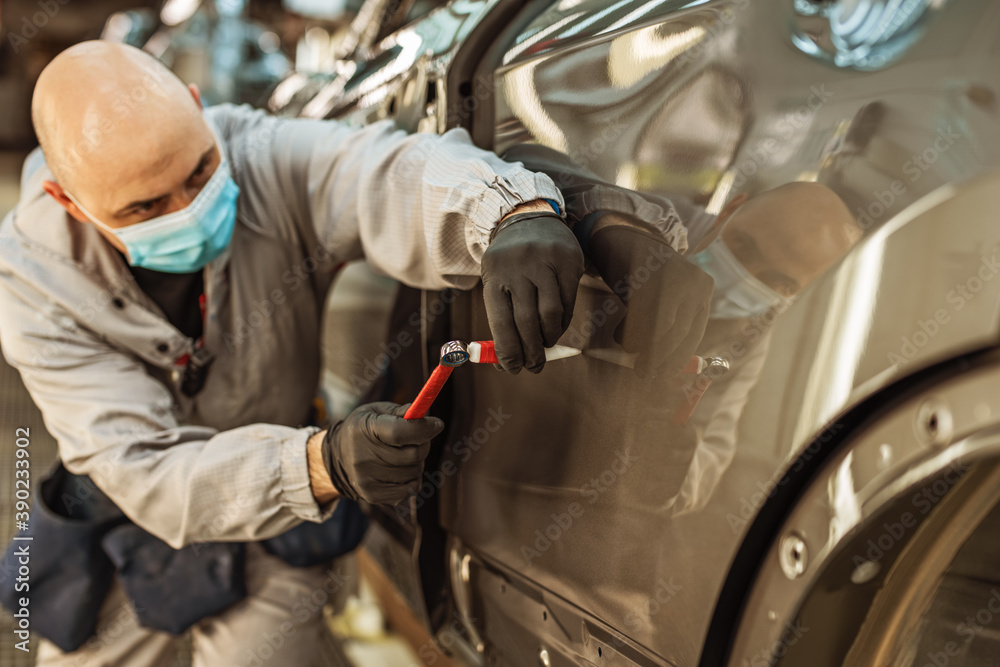 A car factory worker wearing a protective medical mask removes a metal defect with a hand tool. A car factory worker wearing a protective medical mask removes a metal defect with a hand tool.