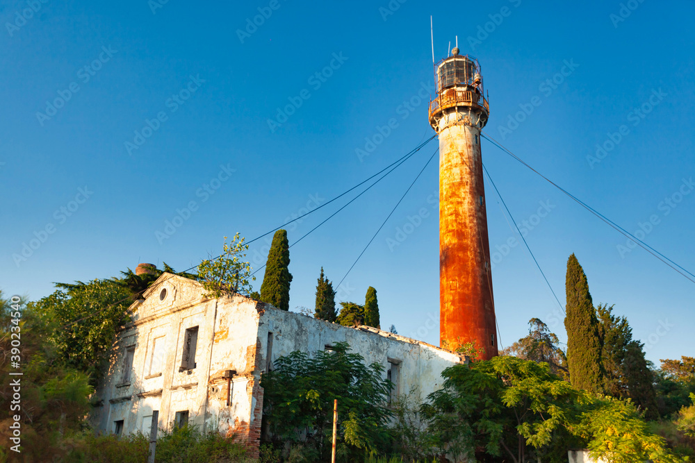 An old rusty lighthouse against a clear blue sky, in the foreground an old building covered with vegetation