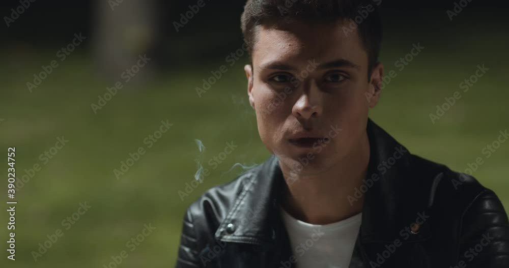 Close up portrait of young thoughtful man smoking alone, sitting at empty park late in evening, looking at camera