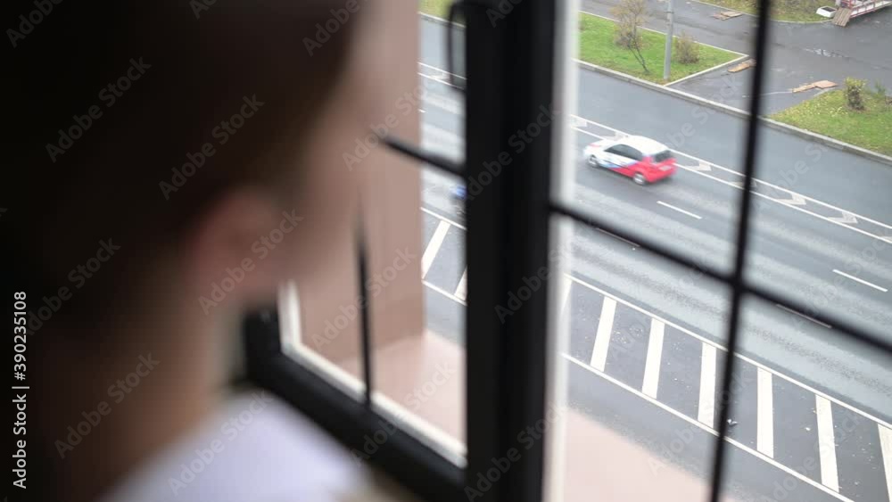 A young woman looks out of the window at passing cars on the road near her house. View of the road with cars from the window of a city house
