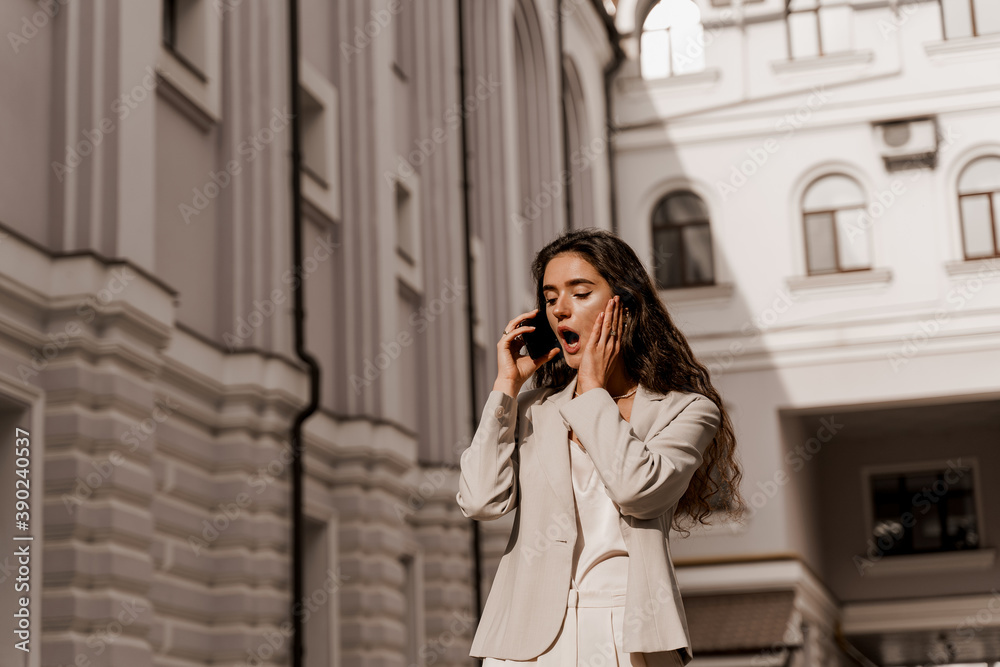 Business woman talking by phone in the city. Communication with friends. Girl with curly hair weared white formal clothes Business woman talking by phone in the city. Communication with friends. Girl with curly hair weared white formal clothes