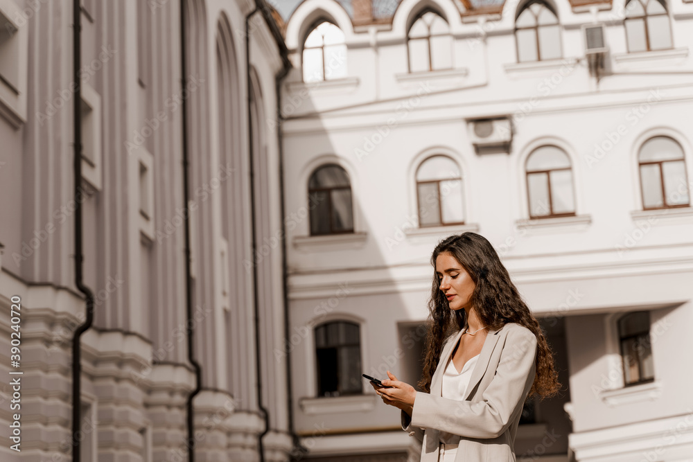 Attractive girl with curly hair holds phone and answers on messages in social networks in the city. Girl is serfing on the internet using smartphone. Manager works on-line by phone.