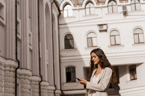 Attractive girl with curly hair holds phone and answers on messages in social networks in the city. Girl is serfing on the internet using smartphone. Manager works on-line by phone.