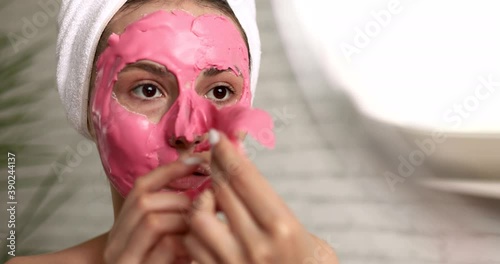 Happy young woman with towel on head taking off pink alginate mask from face while looking at round mirror. Attractive lady using professional cosmetics regularly at home.